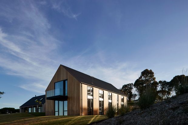 Exterior view of a contemporary timber-clad, single-storey gable-roofed building on a landscaped sloping site. The facade combines vertical warm timber cladding with extensive dark-framed glazing, a projecting glazed balcony, and a dark pitched roof. A smaller matching building is visible behind, with mature trees and open sky surrounding the property.