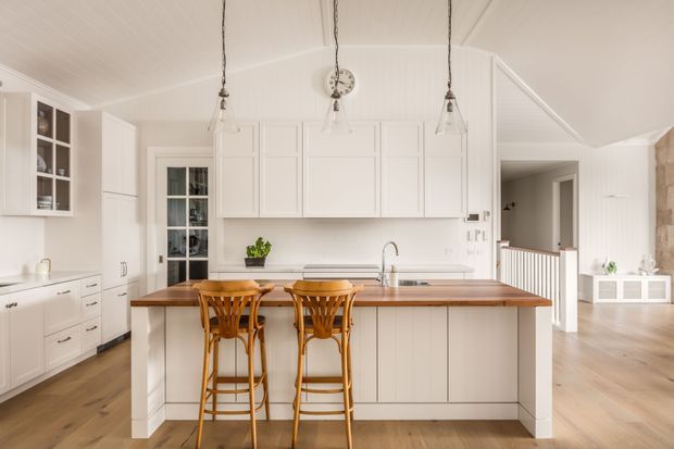 Bright, refined kitchen with a large central island featuring a warm timber benchtop, white paneled cabinetry, integrated sink, and two timber bar stools. The room has white vertical wall panelling, light timber flooring, glass pendant lights, open shelving, and a connected hallway or living area visible beyond the kitchen.