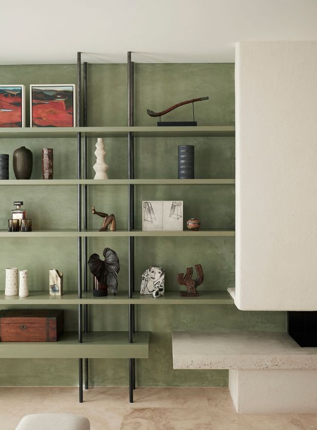 Interior detail of a refined contemporary living space featuring a full-height open shelving system with dark metal uprights and muted green shelves against a textured sage-green wall. The shelves display ceramics, sculptures, artwork, books and decorative objects. At right is a substantial white plaster or stone fireplace volume with a projecting hearth, while the lower portion shows a pale natural stone or concrete floor and a partial upholstered seat.