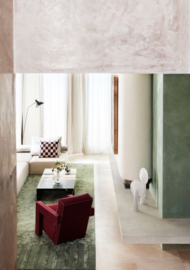 A refined contemporary living room with a cream sectional sofa, green area rug, low marble or stone coffee table, and a deep burgundy accent armchair. Floor-to-ceiling sheer curtains bring in soft daylight, while rounded architectural forms, pale plaster surfaces, timber detailing, and a large muted green feature wall create a calm, tactile interior. A sculptural striped object and checkerboard cushion add small graphic accents.