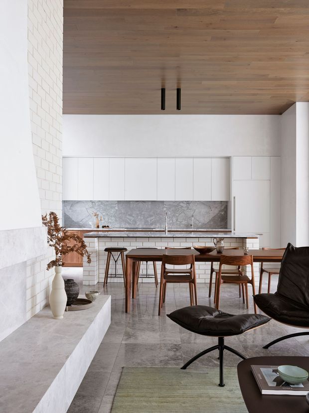 A refined open-plan kitchen and dining area with a minimalist white fit-out, marble-look backsplash, long timber island, and integrated white cabinetry. A warm timber ceiling contrasts with pale stone or concrete flooring and white brick wall surfaces. The dining table and chairs sit beside a black leather lounge chair and ottoman, creating a calm, contemporary living environment.