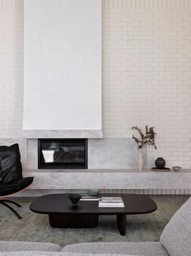 A refined contemporary living room centred on a linear fireplace set into a pale stone-clad mantel and hearth. The backdrop combines a soft white painted brick wall with a tall smooth white chimney breast. A dark low-profile coffee table, sculptural black lounge chair, pale upholstered seating, muted rug, and restrained decorative objects create a calm, sophisticated composition.