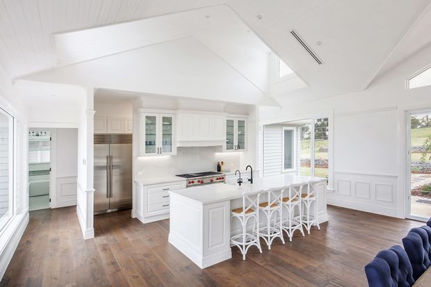 Bright, spacious white kitchen with a large central island, integrated seating, shaker-style cabinetry, stainless steel appliances, and a gas range. The room features warm timber flooring, white paneled walls, a vaulted ceiling with skylight, and abundant glazing overlooking the outdoors. A small portion of a blue upholstered sofa is visible at the edge of the frame.