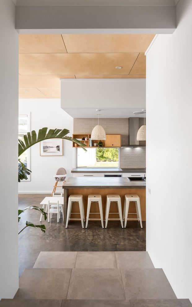 Bright contemporary open-plan kitchen viewed from an adjoining entry or hallway. The kitchen features a large island with four white stools, timber cabinetry, a pale tiled splashback, woven pendant lights, integrated appliances, and a light timber-panelled ceiling. A dining area and indoor plants are visible to the left, with large-format floor tiles in the foreground transitioning to a darker concrete-look kitchen floor.