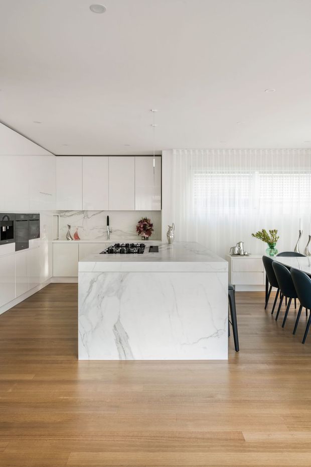 Bright, spacious open-plan kitchen and dining area with a large white marble-look island, integrated white cabinetry, marble-veined splashback, black appliances and fixtures, and warm timber flooring. Sheer curtains diffuse daylight over a dining table with dark upholstered chairs. The overall design is clean, refined, and contemporary.