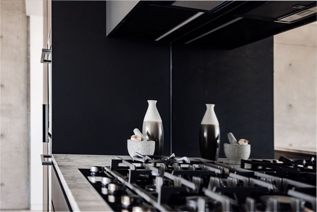 Close-up of a sophisticated contemporary kitchen cooktop beneath a black rangehood. The space features a dark matte splashback and cabinetry or wall surfaces, a pale stone benchtop, a large black gas cooktop, and exposed concrete side walls. Decorative ceramic vessels sit against the dark backdrop.