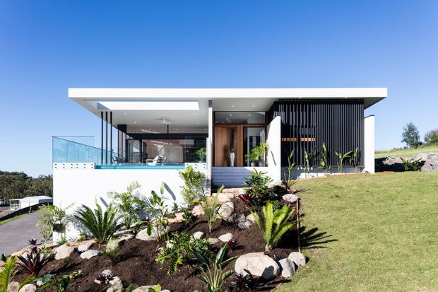 A low, contemporary flat-roofed house set into a landscaped sloping site. The exterior combines crisp white rendered walls, extensive glazing, a glass balustrade, warm timber detailing, and a prominent dark vertical-screen facade. Tropical and native-style planting with rocks fills the foreground garden, while an open terrace and interior spaces are visible through the glazing.