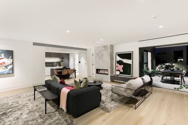 A bright, contemporary open-plan living room with pale timber flooring, a large textured rug, dark modular sofa, sculptural lounge chairs, and minimalist coffee table. The room features recessed lighting, a marble-look feature wall with a linear fireplace, abstract artwork, and extensive opening to an outdoor dining terrace. A modern kitchen is visible beyond the living area.