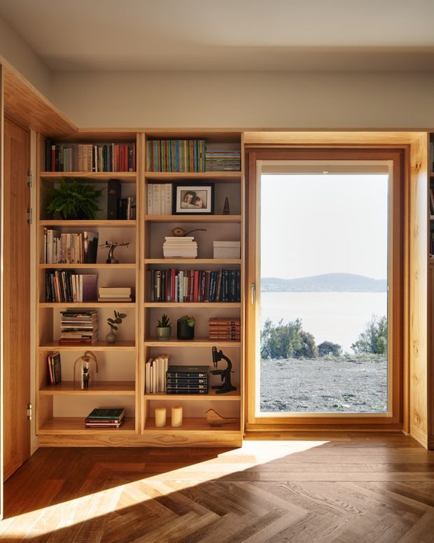 Warm, well-crafted built-in timber bookcases frame a large timber-framed window overlooking a bright coastal or lakeside landscape. The shelving is filled with books and small decorative objects, while sunlight falls across the rich herringbone timber floor. The space has a calm, natural, traditional-meets-contemporary character.