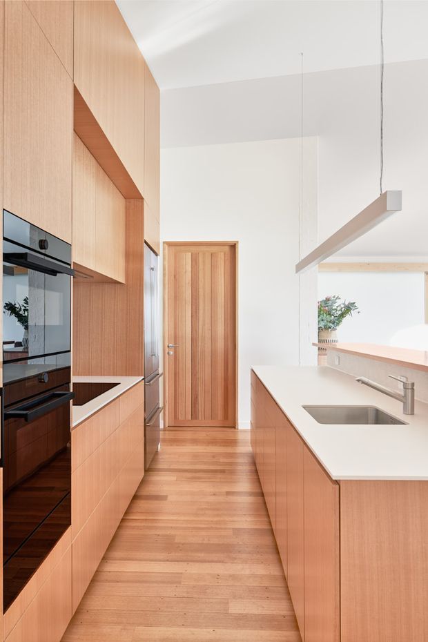 A narrow contemporary kitchen with extensive warm timber cabinetry, integrated black appliances, a white engineered-stone island benchtop and stainless-steel sink. Timber flooring runs through the space toward a vertical timber internal door. A slim linear pendant hangs above the island, and a bright opening with greenery is visible beyond the kitchen.