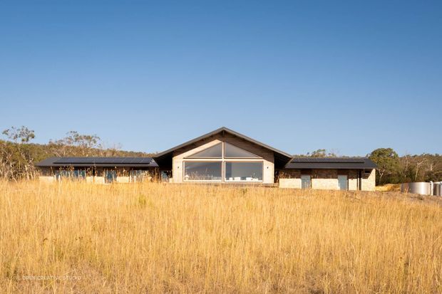 Single-storey contemporary rural home set behind tall golden grasses. The broad low-profile building has dark pitched roof wings and a central gabled pavilion with a large triangular clerestory and full-height glazing. Neutral masonry walls, dark roof cladding, and expansive glazing create a restrained connection to the surrounding landscape.