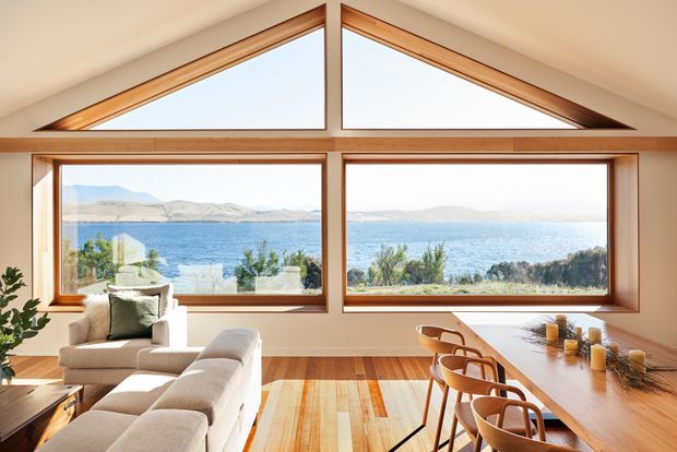 Bright open-plan living and dining space with expansive timber-framed glazing overlooking a lake or broad coastal waterway. The room features a sloped ceiling with distinctive triangular clerestory windows, warm timber flooring and window reveals, a light upholstered sofa, and a long timber dining table with curved wooden chairs. The setting feels airy, warm, and strongly connected to the landscape.