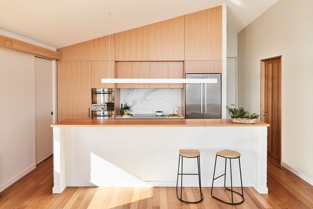 Bright contemporary kitchen with a large white island and timber benchtop, two black-framed stools, extensive light timber vertical-panel cabinetry, integrated stainless-steel appliances, and a marble-look splashback. Warm timber flooring and a timber-framed doorway add warmth to the clean, open design.