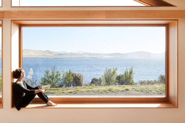 Interior view centered on a very large picture window with a deep timber surround and horizontal timber wall detailing. A person sits on the broad window sill, looking out across a lake toward low, dry hills. The room is bright and calm, with strong natural light and an expansive framed landscape view.