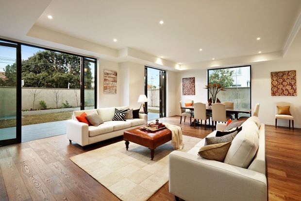 A bright, spacious open-plan living and dining area with warm timber flooring, cream walls, recessed downlighting, and extensive black-framed floor-to-ceiling glazing. Two cream sofas face a central leather ottoman and rug, while a dining table and chairs sit beside the windows. The glazing opens toward a small landscaped courtyard.