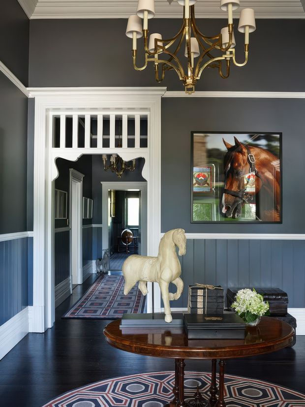A refined traditional hallway or entry space with dark blue-grey walls, white decorative trim and wainscoting, a highly detailed white coffered ceiling, and dark timber flooring. A brass and cream multi-arm chandelier hangs prominently overhead. The room features a polished oval timber console table with books, a floral arrangement and a sculptural rocking horse, while a large framed horse portrait and patterned runner rug add character. An ornate doorway with transom glazing leads into a long corridor with repeated trim and another chandelier.