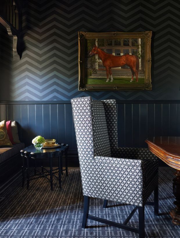 A moody, richly patterned sitting room with dark navy-blue vertical wall panelling below grey-and-black chevron wallpaper. A tall geometric-patterned wingback chair dominates the foreground beside a small round black side table, while part of a striped sofa and polished dark wood table are visible. An ornate framed horse painting hangs above the panelling, and patterned carpet covers the floor. The room has dramatic lighting and a sophisticated, traditional club-like atmosphere.