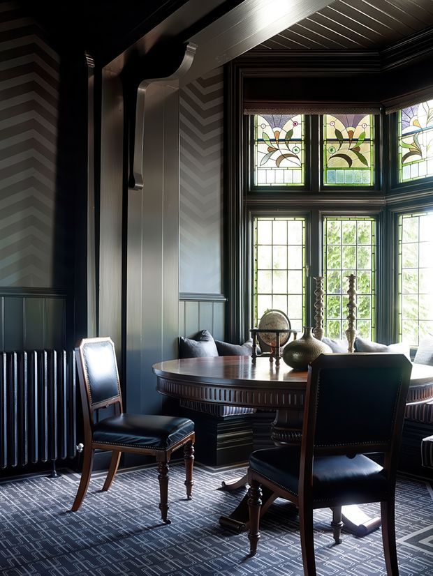 A richly detailed, dark-toned dining room or formal sitting area with a round dark timber table, upholstered timber chairs, decorative objects, and a large bay window. The room features dark vertical wall panelling, patterned chevron wallpaper, ornate trim, a radiator, patterned carpet, and prominent leadlight stained-glass transom windows. Natural daylight creates contrast against the moody interior.