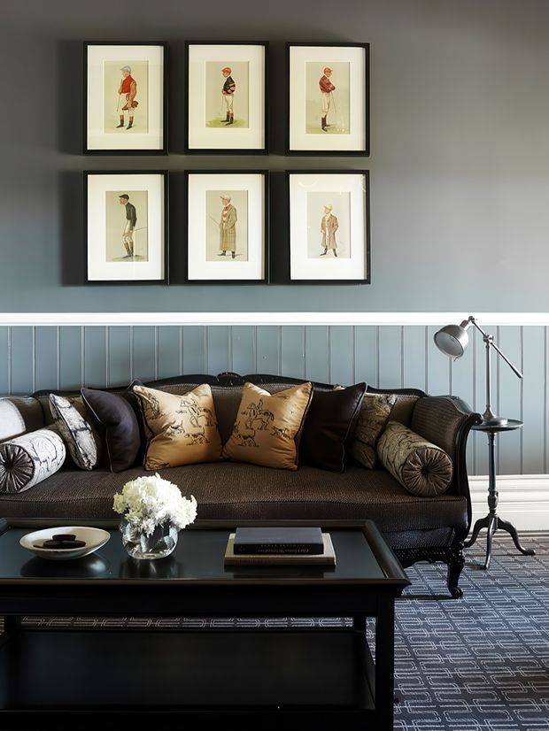 A refined living room with a dark, tailored sofa arranged against blue-grey vertical wall panelling. Six framed vintage-style equestrian prints are arranged in a symmetrical two-row gallery above the sofa. Black side tables, a sculptural floor lamp, patterned cushions, a dark coffee table, floral arrangement and geometric rug create a formal, classic interior.