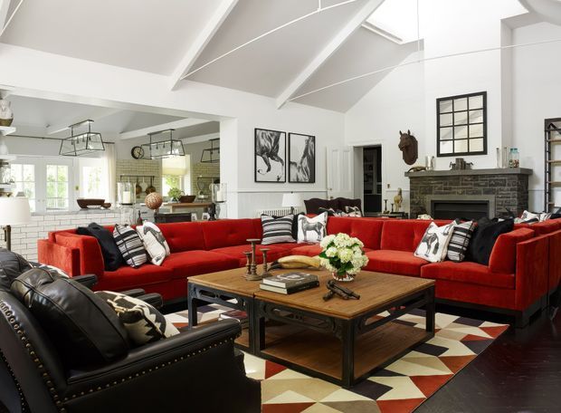 Bright, spacious living room with a dramatic vaulted white ceiling and exposed beams, large red sectional sofas, a black leather armchair, a substantial rustic wood-and-metal coffee table, and a geometric area rug. The room includes a dark masonry fireplace with mantel, black-framed interior window, artwork, shelving, and an open connection to a light-filled kitchen or dining area. The styling combines traditional architectural elements with bold contemporary furnishings and industrial accents.