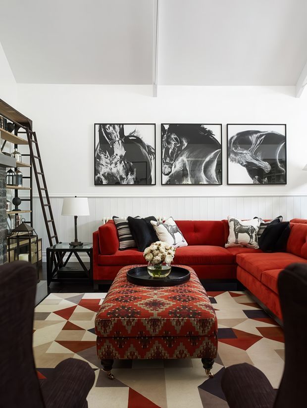 A stylish living room with a vivid red L-shaped sectional sofa, patterned red upholstered ottoman, geometric area rug, black-and-white animal artwork, and open industrial-style shelving. White vertical wall panelling and a white vaulted ceiling create a bright backdrop, while dark timber and metal accents add contrast.