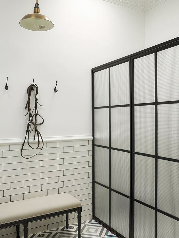 A refined bathroom or dressing-room corner with white subway-tiled lower walls, white painted upper walls, ornate ceiling cornice, black-framed textured-glass partition doors, and a geometric patterned floor. A dark vintage-style bench sits beneath wall-mounted hooks holding leather straps, while a brass pendant light adds warmth. The scheme combines traditional detailing with industrial-inspired metal and glass.