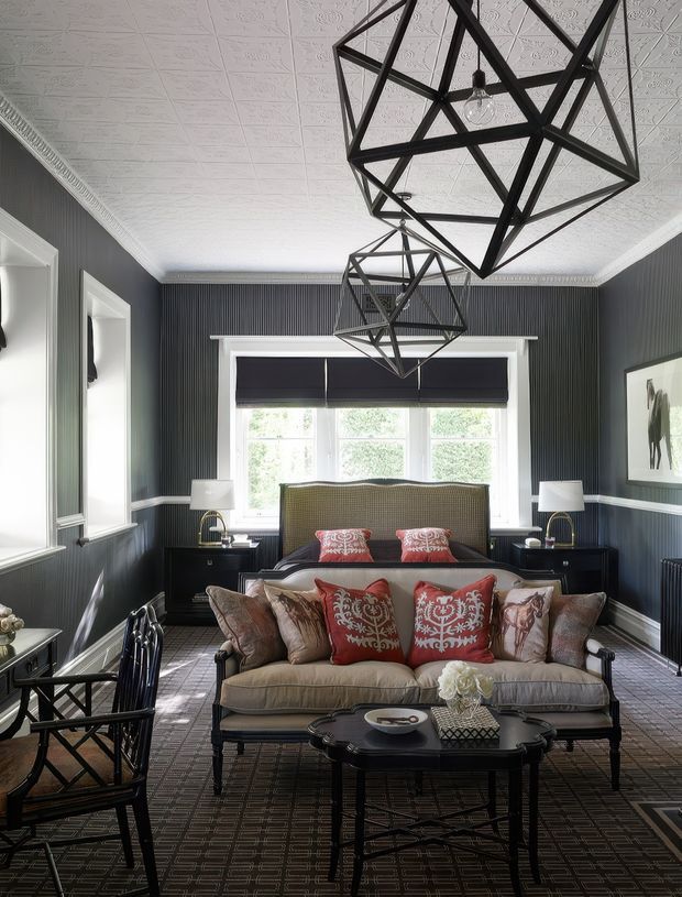 A refined bedroom sitting area with a four-poster or upholstered bed positioned behind a large sofa, dark vertically panelled walls, patterned carpet, ornate pressed-tin ceiling, white decorative cornices, and large geometric black metal pendant lights. Red and neutral patterned cushions add contrast, while tall windows provide natural light. The room has a layered, formal and eclectic character.