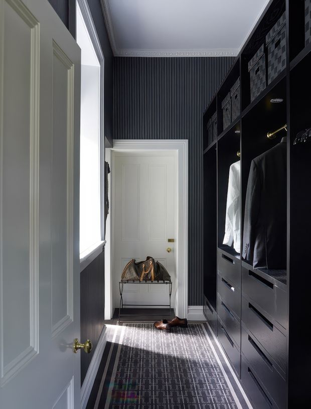 A narrow, elegant walk-in wardrobe or dressing room viewed from an open paneled door. One wall has tall dark built-in cabinetry with hanging clothes, drawers, shelves, and storage boxes. The opposite wall is dark vertically fluted paneling with a large window, while white paneled doors and ornate ceiling cornice provide contrast. A patterned runner extends along the floor, with a small metal bench or luggage stand near the far door and shoes on the floor.