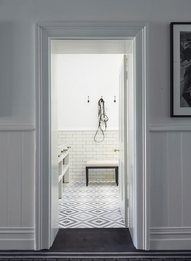 View through a paneled doorway into a bright, compact bathroom or dressing area. The room features white subway-tile wall cladding, a bold grey-and-white geometric patterned tile floor, a dark upholstered bench, white cabinetry or shelving, and traditional layered door and wall mouldings in a grey-painted interior. A framed monochrome artwork is visible on the surrounding wall.