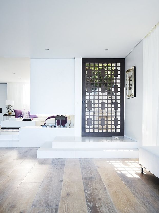 Bright minimalist interior showing a white open-plan living area and entryway. Pale wide-plank timber flooring leads to a raised platform and a dramatic black gridded front door with glazed openings. White built-in cabinetry and walls dominate, with a glimpse of a sofa and purple cushions in the living room beyond.