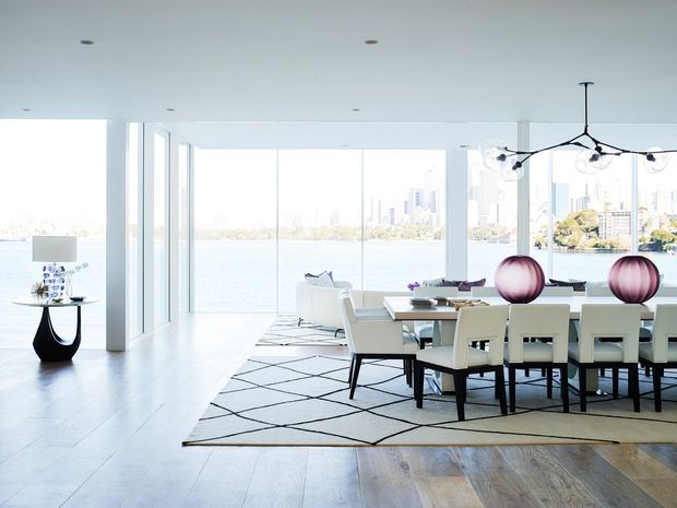 Bright, spacious contemporary open-plan dining and living area with floor-to-ceiling glazing overlooking a waterfront city skyline. The room features pale timber flooring, a large geometric area rug, a long dining table with upholstered chairs, sculptural pendant lighting, and minimalist furnishings.