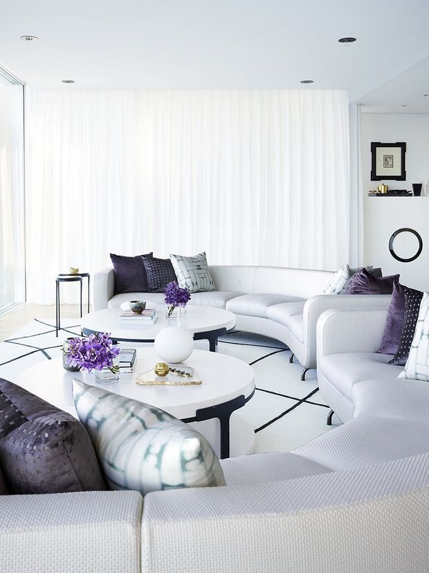 Bright, sophisticated living room with multiple white upholstered sofas and rounded coffee tables arranged over a white rug with bold black geometric lines. Sheer floor-to-ceiling curtains diffuse abundant daylight, while purple and patterned cushions and floral arrangements provide restrained accents. A minimalist white wall unit with framed art, shelving, and a circular decorative feature completes the refined contemporary setting.