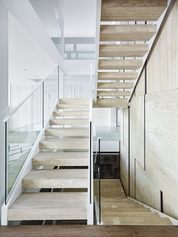 A bright, minimalist multi-level stairway with floating pale stone or concrete treads, slim black handrails, and clear glass balustrades. The stairs wrap through a tall open void beside a textured light stone feature wall, with white walls, glazing, and glimpses of timber flooring below.
