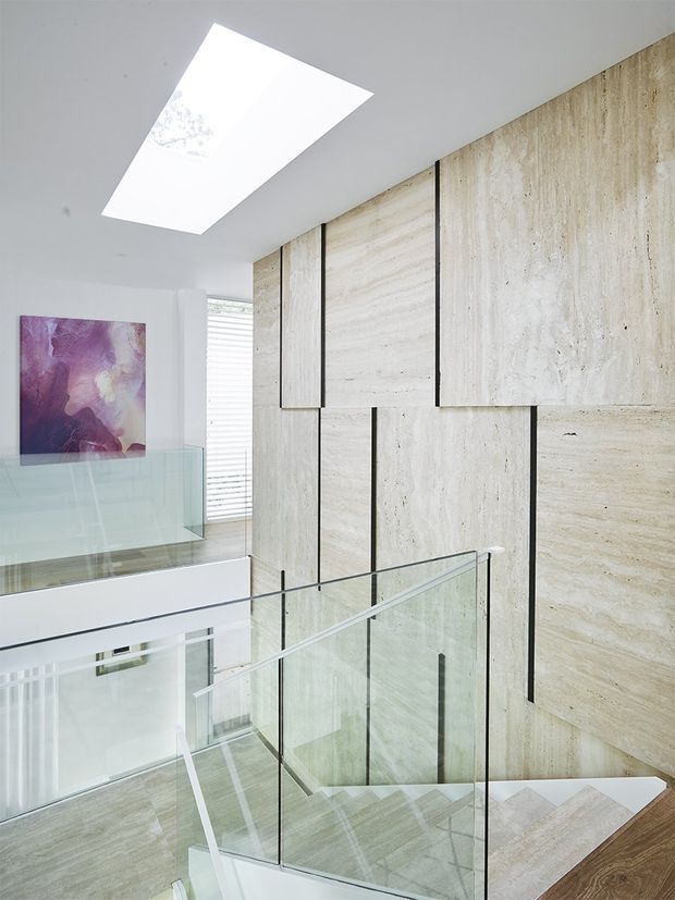 A bright contemporary stairway and landing with a glass balustrade, pale stone or travertine wall panels divided by dark vertical joints, and matching light stone stair treads. A large rectangular skylight illuminates the space, with white walls, a glass guardrail, and an abstract purple artwork visible on the adjacent landing.