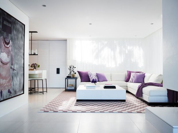 Bright, spacious contemporary living room with a large white sectional sofa, oversized white rectangular coffee table, sheer floor-to-ceiling curtains, pale tiled flooring, and a purple, cream, and grey checkered area rug. A compact bar or kitchen zone is visible at the rear, while large abstract artwork and sculptural accessories add visual interest.