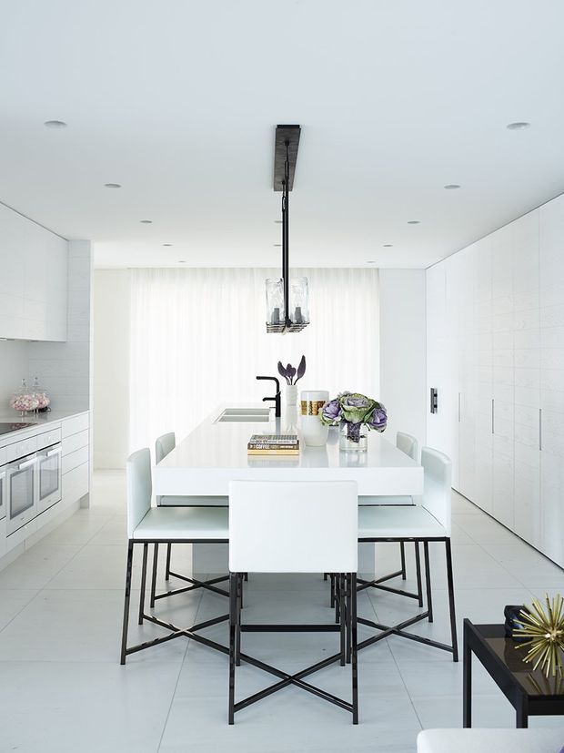 Bright, predominantly white open-plan kitchen and dining area with a long white island or dining table, white upholstered chairs on black metal frames, integrated white cabinetry, tiled walls and flooring, and a contemporary linear pendant light. A built-in oven and cooktop are visible along the left wall, while sheer curtains provide soft natural light at the rear.