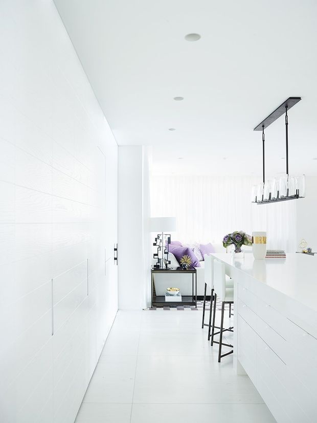 Bright, predominantly white open-plan interior showing a sleek kitchen island with white horizontal-panel cabinetry, a black-framed linear pendant light, and black bar stools. The space opens toward a living area with a purple sofa, sheer curtains, a small black console, and decorative accents. Recessed ceiling lights and pale large-format flooring reinforce the clean contemporary aesthetic.