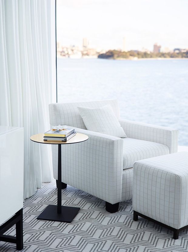 A bright, refined waterfront sitting area with a white grid-upholstered armchair and matching ottoman beside a small round side table. Sheer white curtains frame expansive glazing with a calm body of water and distant urban shoreline beyond. A bold geometric black-and-white rug anchors the seating arrangement.