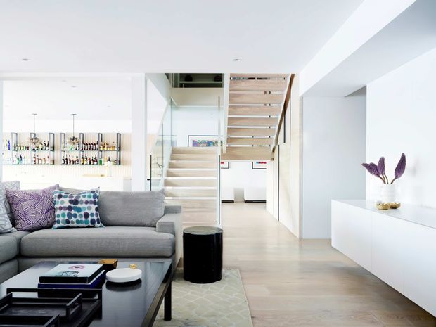 Bright, open-plan contemporary living area with a large grey sectional sofa, patterned cushions, black coffee table, pale timber flooring, white built-in cabinetry, and a sculptural open-tread staircase. A compact home bar with backlit bottle shelving is visible in the background, while a corridor and artwork extend beyond the stair.