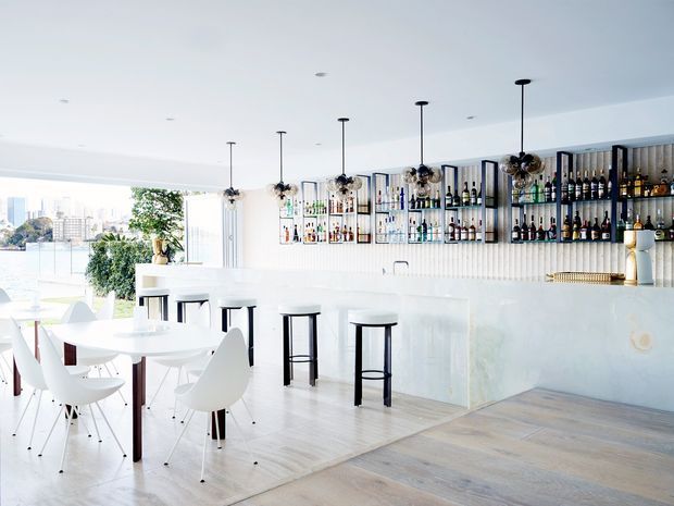 Bright, spacious open-plan home bar and dining area with a long white stone bar counter, black stools, pendant lighting, and wall-mounted open bottle shelving. A white dining table with modern sculptural chairs sits in the foreground, while large glazing opens to an outdoor view. The space has pale timber flooring, white walls and cabinetry, and a clean contemporary aesthetic.