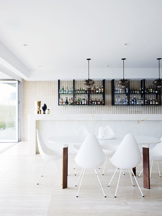 Bright, minimalist dining and home bar area with a long white dining table surrounded by sculptural white chairs. A built-in dark-framed liquor display with glass shelves is set into a vertically textured light wall above a white counter or sideboard. Black spherical pendant lights hang over the table, while large sliding glazing at left brings in abundant daylight. The space has a refined, monochromatic contemporary aesthetic with warm timber chair legs and pale flooring.