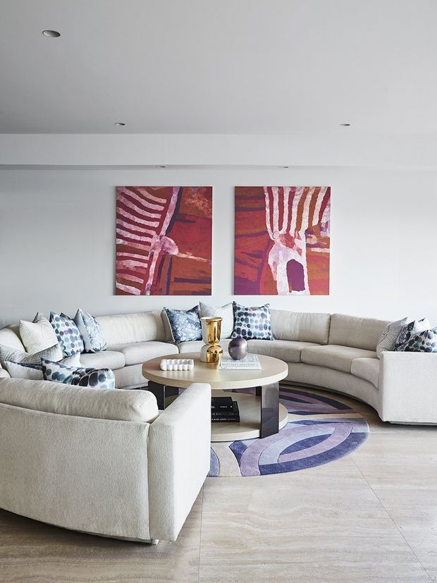 Bright, spacious contemporary living room with a large cream sectional sofa and matching armchair arranged around a round coffee table. The room has pale stone-look flooring, white walls and ceiling with recessed downlights, colourful abstract artwork, patterned cushions, and a circular purple, blue and cream rug.