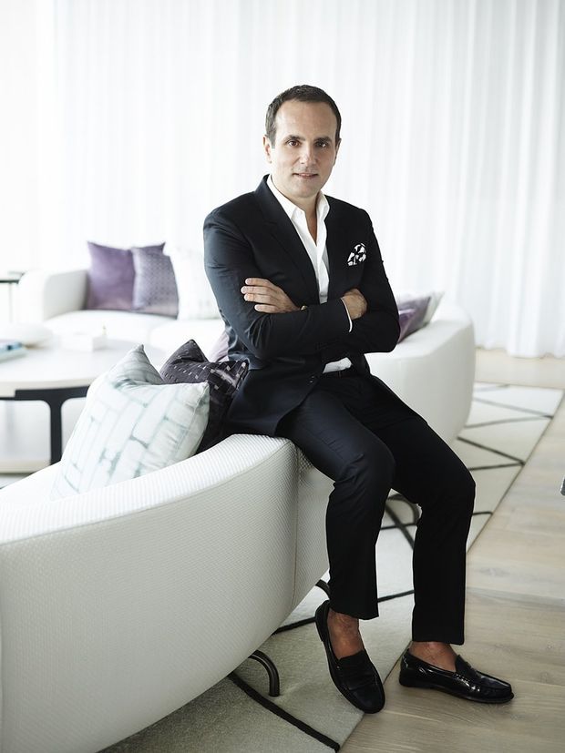 A portrait of a man in a black suit seated on a white curved sofa in a bright, refined living room. The room features sheer white curtains, pale timber flooring, a light area rug with dark geometric lines, a white coffee table, and neutral upholstered seating with muted purple and patterned cushions.