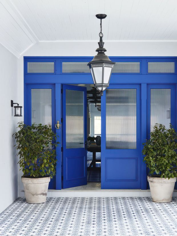 A bright, formal entryway with vivid cobalt-blue glazed double doors and matching transom windows, set within white walls beneath a white beadboard ceiling. The entrance features patterned encaustic-style floor tiles, large white planters with leafy greenery, black traditional lantern lighting, and an ornate brass door knocker. The open doors reveal a glimpse of the interior beyond.