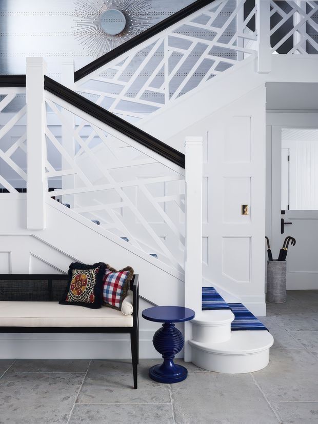 Bright, highly detailed stair hall with white geometric balustrades and panelled walls, black handrails, and a decorative sunburst wall clock. A white built-in bench with patterned cushions sits beside a glossy navy-blue side table, while curved white stairs descend onto a grey stone floor. The space has a crisp, graphic, coastal-traditional character with strong contrast and layered architectural detailing.