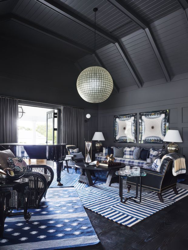 A dark, highly styled living room with a dramatic pitched, exposed-beam ceiling and charcoal wall panelling. The room features a glossy black grand piano, black leather seating, a blue-and-white plaid sofa, patterned blue rugs, woven lounge chairs, mirrored artwork, table lamps, and a large spherical pendant light. Bright glazing and an open door connect the room to the outdoors.