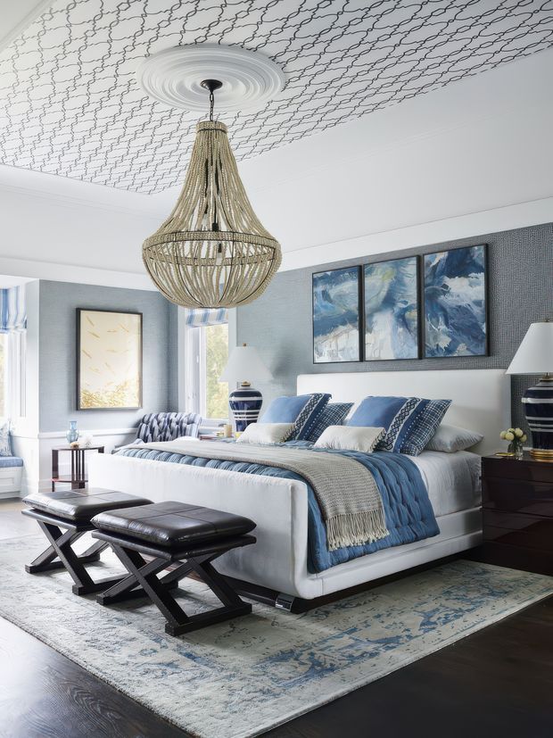 A bright, refined bedroom with a large upholstered white bed dressed in layered blue and white bedding, patterned cushions, and a fringed throw. The room features blue-grey textured wall covering, dark timber flooring, a large pale patterned rug, black leather X-benches at the foot of the bed, dark bedside furniture, blue-and-white ceramic lamps, framed artwork, and an oversized woven pendant light. A decorative repeating pattern covers the ceiling around a central medallion.
