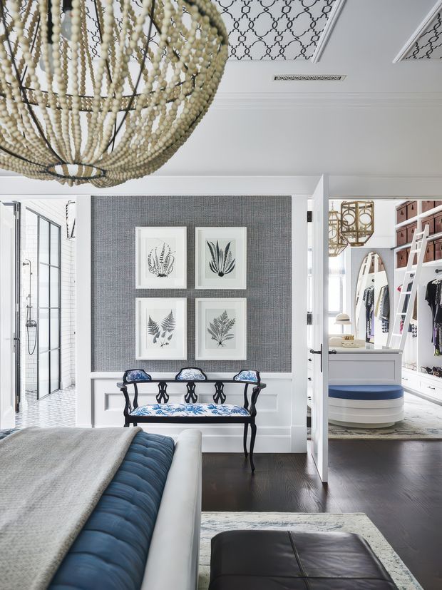 A refined bedroom interior with a large cream bead chandelier, dark timber flooring, a blue upholstered bed, and a decorative bench beneath four botanical prints. The feature wall combines grey textured wallpaper with white wainscoting. An open doorway reveals a bright walk-in wardrobe and a tiled bathroom beyond. The ceiling has patterned inset panels and recessed linear vents, contributing to an elegant, tailored aesthetic.
