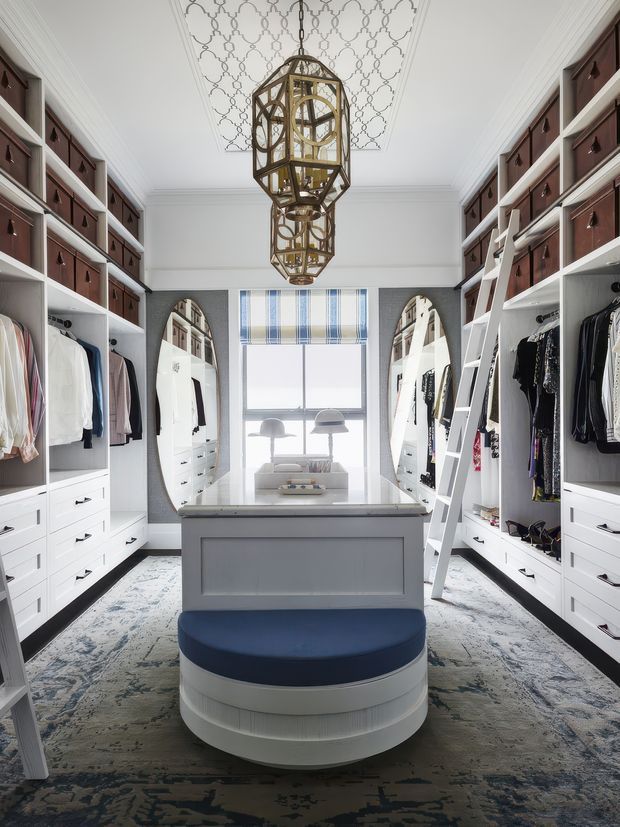 A bright, spacious walk-in wardrobe with symmetrical white built-in cabinetry, open shelving, drawers, hanging clothes, full-height mirrors, and leaning ladders. A central island with a grey benchtop and a round upholstered blue seat anchors the room. The space features a patterned carpet, white architectural mouldings, a decorative patterned ceiling, and an ornate bronze-and-glass pendant chandelier.