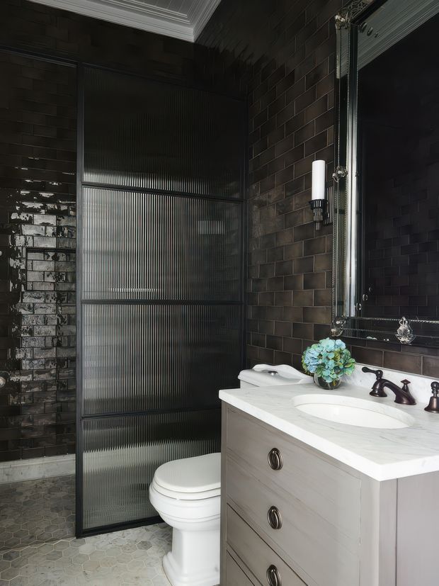 A dramatic, compact bathroom with dark glossy subway-tile walls, a fluted translucent glass shower enclosure, and a white traditional vanity with a pale stone countertop and integrated basin. A white toilet, dark vintage-style tapware, ornate mirror framing, wall sconces, and a small blue floral arrangement complete the space. The room has a moody, refined character with contrasting light fixtures and fittings.